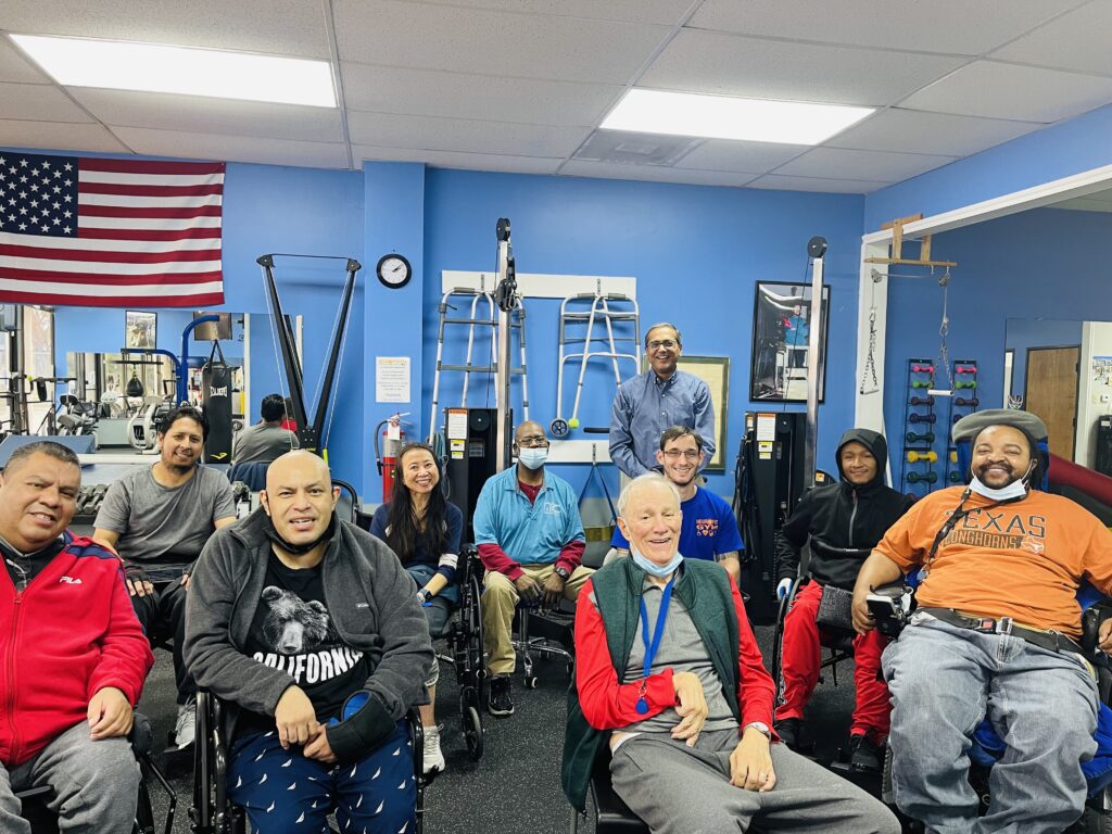 A group of elderly individuals seated in a community room with exercise equipment and an American flag.